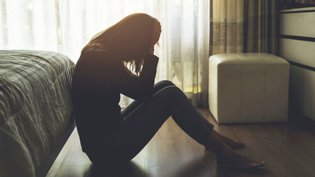 Woman sitting on bedroom floor struggling with depression, representing mental health challenges and the need for support at San Diego Wellness Center.