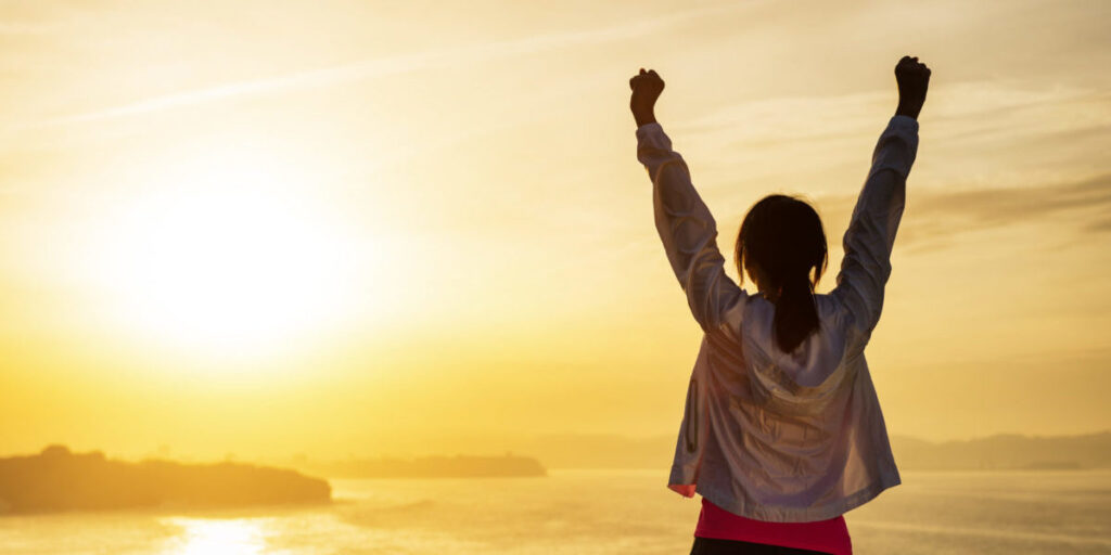 Person raising arms in celebration at sunrise by the ocean, symbolizing hope and recovery at San Diego Wellness Center.