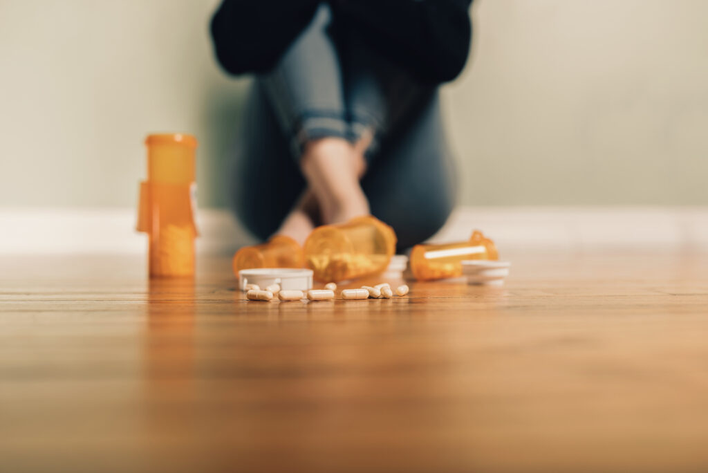 Prescription pill bottles and tablets on the floor with a person sitting in the background, representing anxiety and addiction.